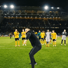 a man in a black hoodie stands on a soccer field in front of a group of players in yellow jerseys