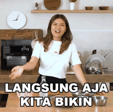 a woman in a white shirt is standing in a kitchen with the words langsung aja kita bikin written above her