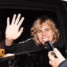 a man with curly blonde hair is waving from a car window .