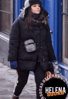 a woman wearing a black jacket and a hat is walking down a street with a helena squad logo in the corner