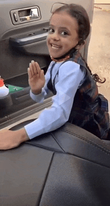 a little girl is sitting in the back seat of a car waving at the camera