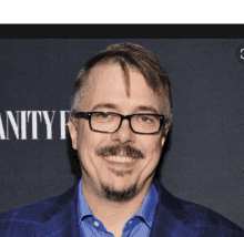 a man wearing glasses and a blue shirt is smiling in front of a vanity fair sign