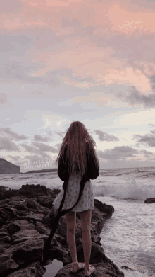 a woman in a white dress stands on a rocky shoreline looking at the ocean