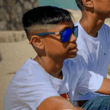 a boy wearing blue sunglasses sits next to another boy wearing a white shirt