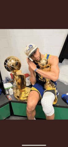 a man in a golden state warriors uniform sits on a bench holding two trophies