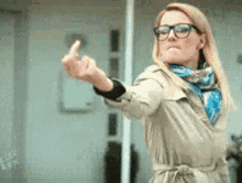 a woman wearing glasses and a trench coat is giving a middle finger .