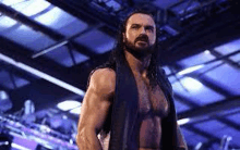 a shirtless wrestler with long hair and a beard is standing in a ring .