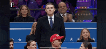 a man in a suit and tie is standing in front of a crowd wearing a red hat that says trump .