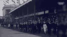 a black and white photo of people walking in front of a stadium with the olympics logo on the bottom