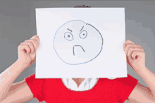 a woman in a red shirt is holding up a paper with an angry face drawn on it