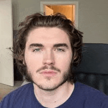 a man with a beard and long hair is wearing a blue shirt and sitting in a chair .