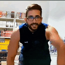 a man with a beard and glasses is standing in front of a bookshelf and pointing at the camera .