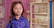 a young girl with a beard and a blue shirt is standing in front of a shelf filled with tiaras .