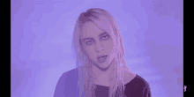 billie eilish is wearing a black shirt and making a funny face in front of a purple background .