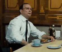 a man in a white shirt and tie is sitting at a table with a plate of food and a cup of coffee