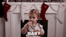 a little girl is eating a cookie in front of a fireplace with stockings hanging on it and the word baby is visible