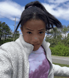 a girl wearing a tie dye shirt and a white jacket takes a selfie