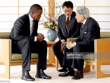 a man in a suit sits on a couch talking to two other men