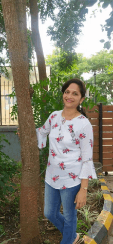 a woman standing next to a tree wearing a white top with red flowers on it