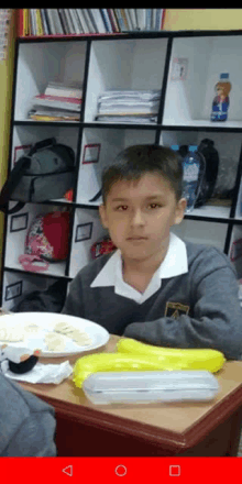 a young boy in a school uniform is sitting at a desk with a plate of food on it .