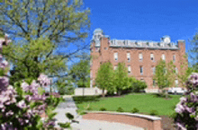 a large brick building with a clock tower is surrounded by trees and flowers on a sunny day .