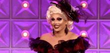 a drag queen wearing a hat and earrings smiles on a purple background