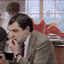 a man in a suit and tie is biting his nails while sitting at a desk in front of a window .