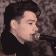 a young man is singing into a microphone in front of a patterned wall .