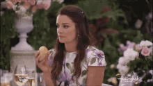 a woman in a floral dress is eating an apple while sitting at a table with flowers in the background ..