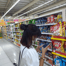 a woman wearing a mask looks at her phone in a grocery store aisle