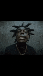 a man with dreadlocks and a gold chain around his neck is standing in front of a wall .