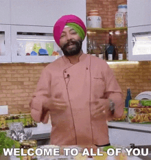 a man in a pink chef 's jacket and turban is standing in a kitchen and says welcome to all of you .