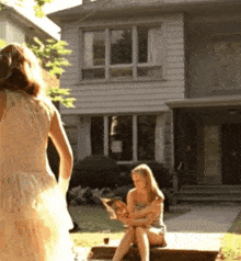 a woman in a white dress is standing in front of a house while another woman sits on the porch reading a magazine