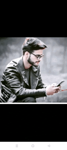 a young man wearing glasses and a leather jacket is looking at his phone