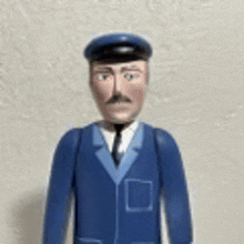 a toy man wearing a blue suit and hat is standing in front of a white wall .