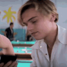 a young man in a white shirt is looking at a cell phone while sitting at a pool table .