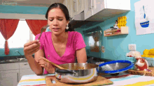 a woman in a pink shirt is holding a fork and spoon in front of a pot in a kitchen