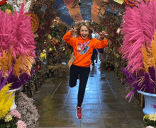 a woman in an orange sweatshirt is jumping in the air in front of flowers