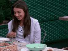 a woman is sitting at a table with a green bowl in front of her and a green fence behind her .