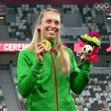 a woman in a green jacket holds a medal and a stuffed animal in front of a sign that says ceremony