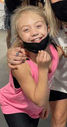 a girl wearing a pink tank top and a black face mask smiles for the camera