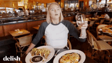 a woman is sitting at a table with plates of food and a glass of wine with the word delish on the bottom right