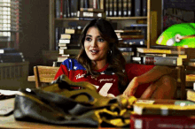 a woman wearing a red shirt with the number 21 on it is sitting at a desk in a library surrounded by books .