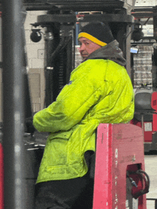 a man wearing a yellow jacket and a black hat sits on a forklift