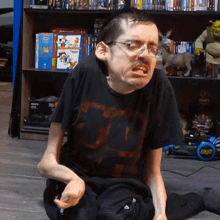 a man wearing glasses and a black shirt with the letter t on it is sitting on the floor in front of a bookshelf