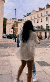 a woman in a white dress is walking down a sidewalk in front of a building that has the letter t on it