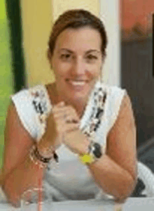 a woman in a white shirt is sitting at a table with her hands folded and smiling .