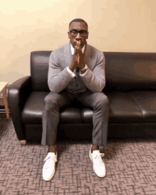 a man sits on a black couch with his hands folded