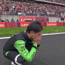 a man in a green and black jacket is kneeling down on a race track covering his face with his hands .