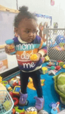 a little girl wearing a sweater that says mom dad me is standing in a room full of toys